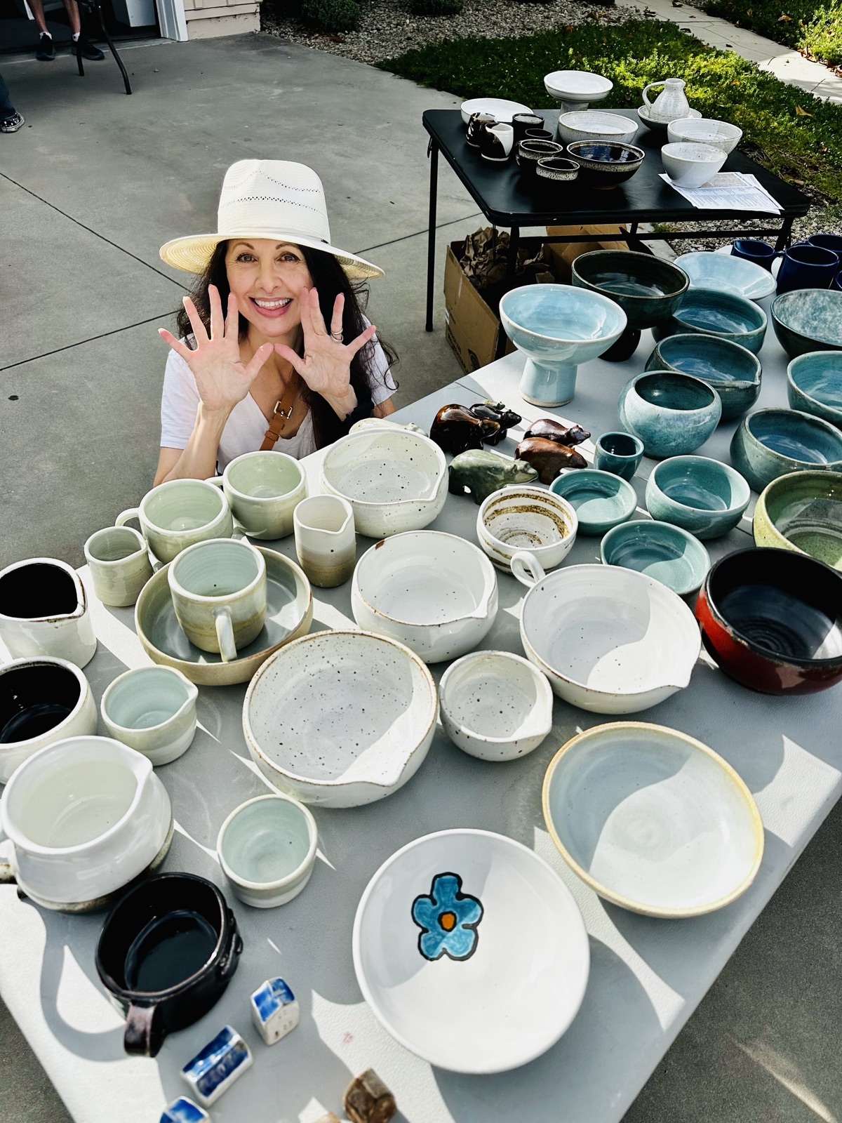 Suzanne Luna in a sun hat behind a table of her pottery, hands raised